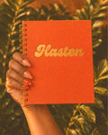 A hand holds a coral-orange spiral notebook with the word 'Hasten' in metallic gold script against a background of green foliage.