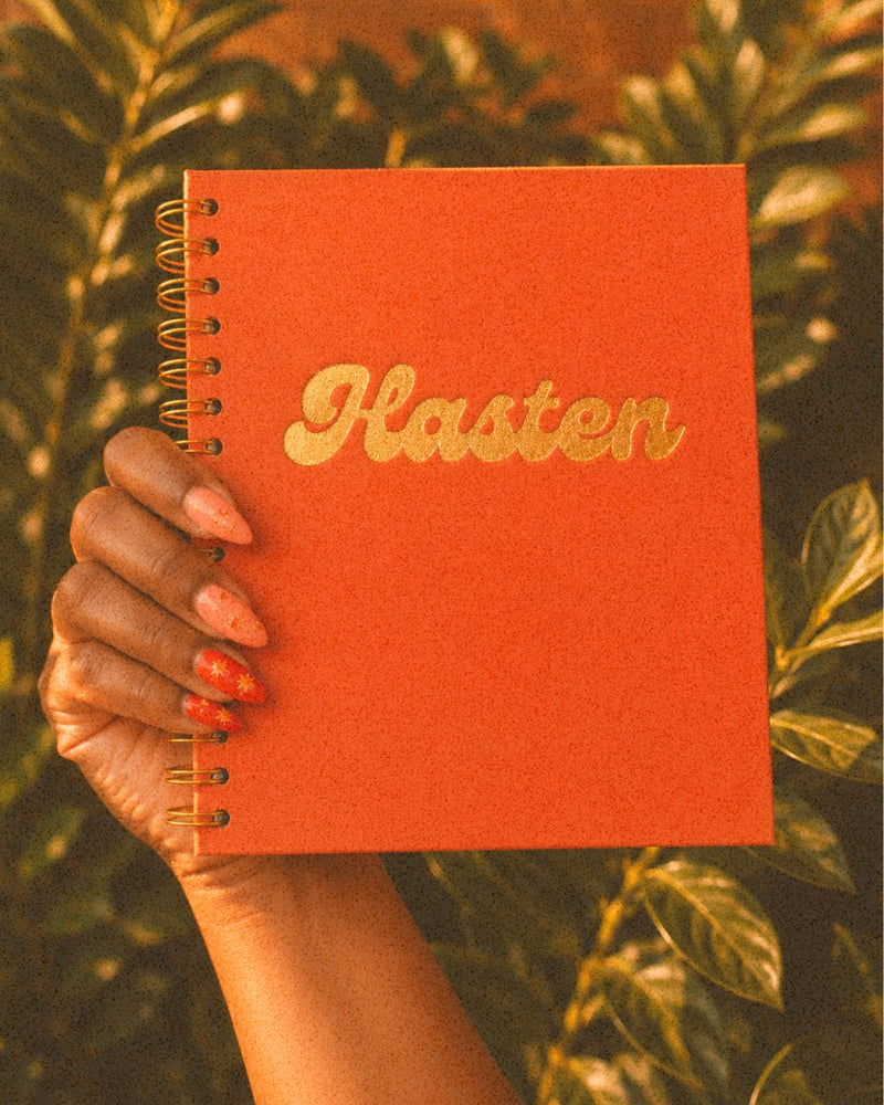 A hand holds a coral-orange spiral notebook with the word 'Hasten' in metallic gold script against a background of green foliage.