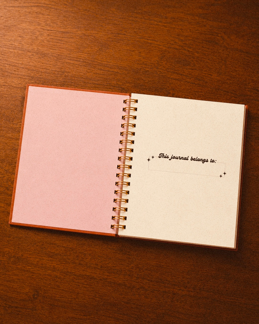 An open spiral-bound journal with a pink left page and an off-white right page, placed on a dark brown wooden surface. The right page has a small handwritten note in script.