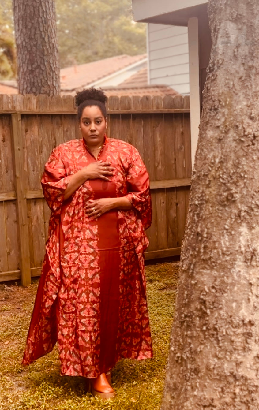 A woman wearing a rust-red patterned kaftan dress stands outdoors in a backyard setting with a wooden fence and a large tree trunk in the foreground.