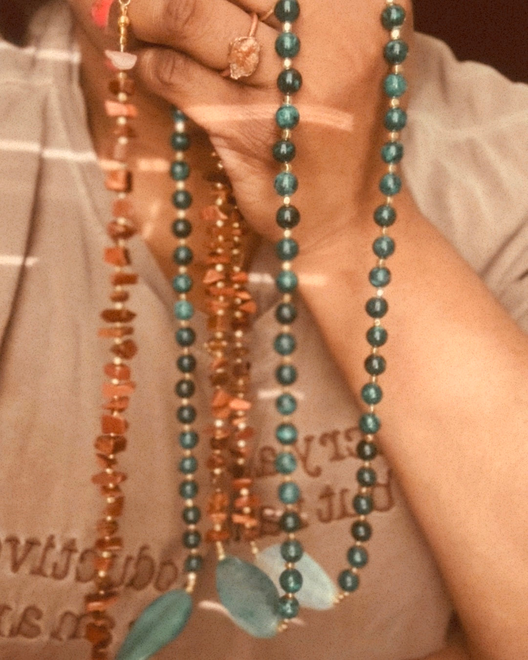 A close-up of a person wearing multiple beaded necklaces, including one with dark teal beads and two with copper-toned beads, over a beige top.