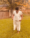 A woman wearing a cream-colored two-piece matching set consisting of a short-sleeve top and wide-leg pants, standing barefoot in a grassy backyard.
