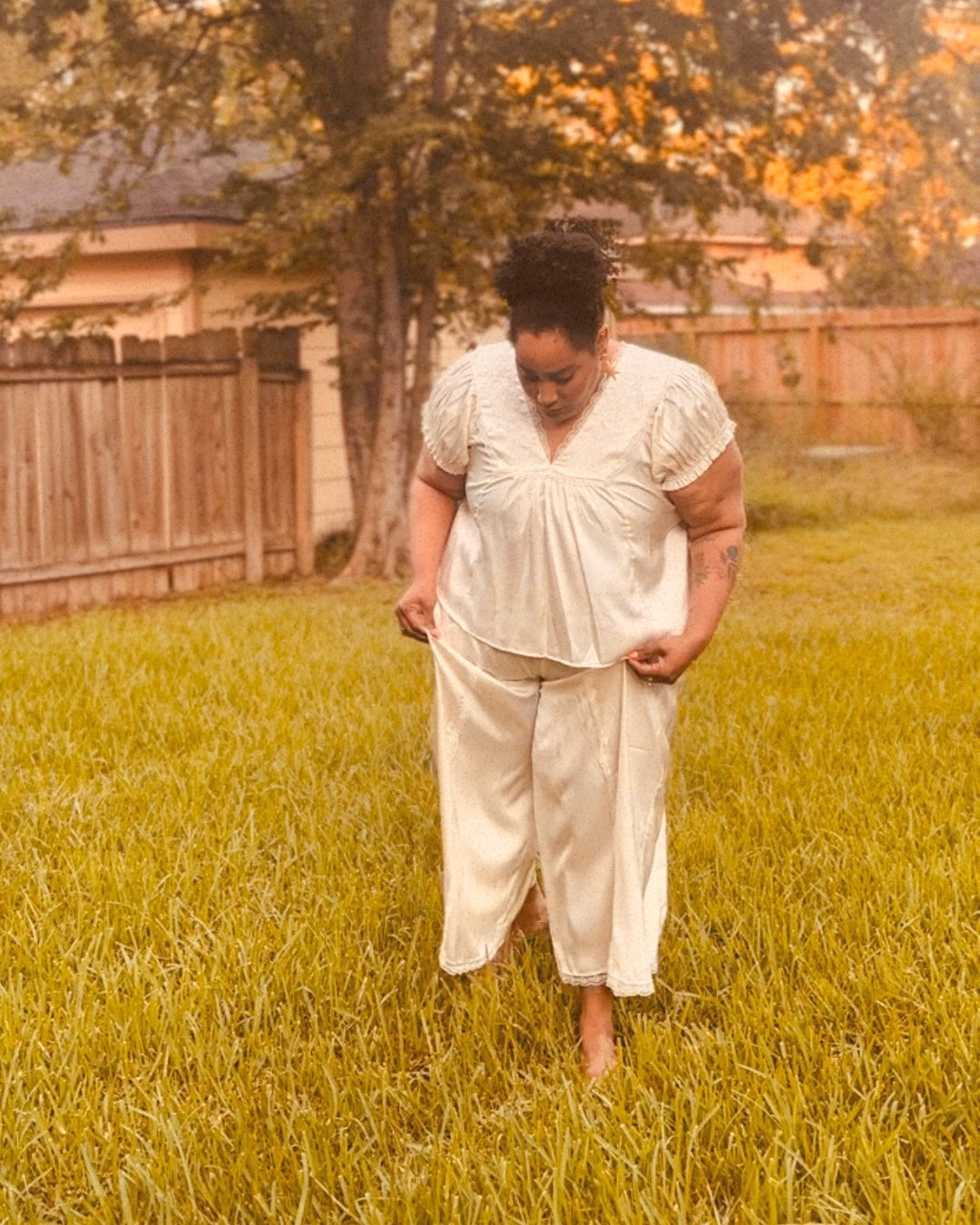 A woman wearing a cream-colored two-piece matching set consisting of a short-sleeve top and wide-leg pants, standing barefoot in a grassy backyard.