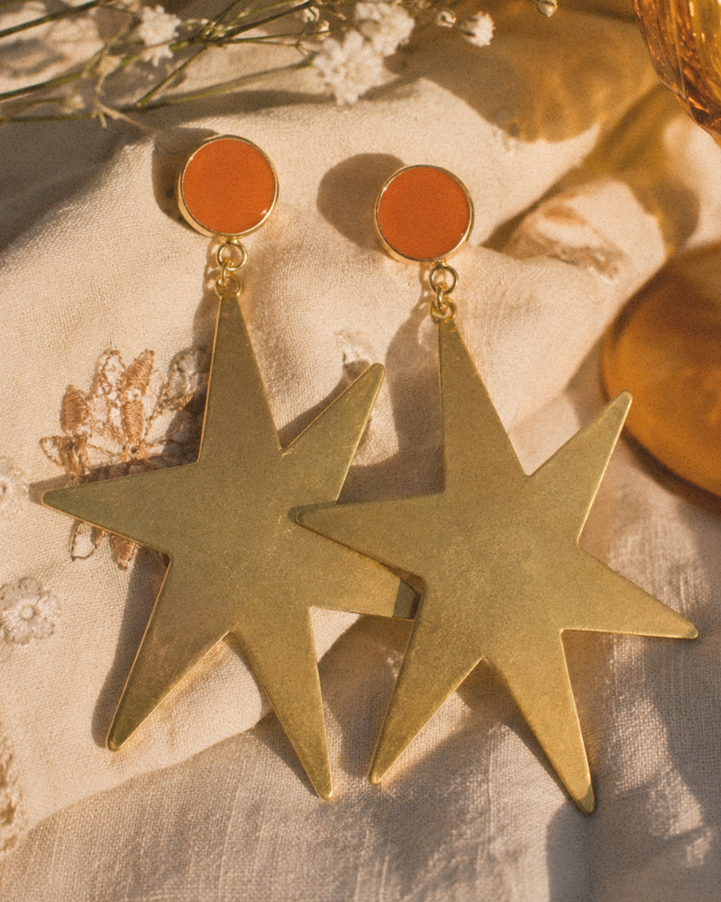 Gold star earrings with orange circular tops on a textured surface.