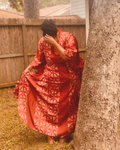 A person outdoors wearing a flowing red or wine colored patterned kaftan garment, standing beside a large tree trunk in a backyard setting with a wooden fence behind them.