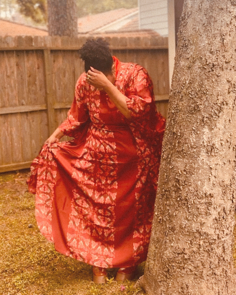 A person outdoors wearing a flowing red or wine colored patterned kaftan garment, standing beside a large tree trunk in a backyard setting with a wooden fence behind them.