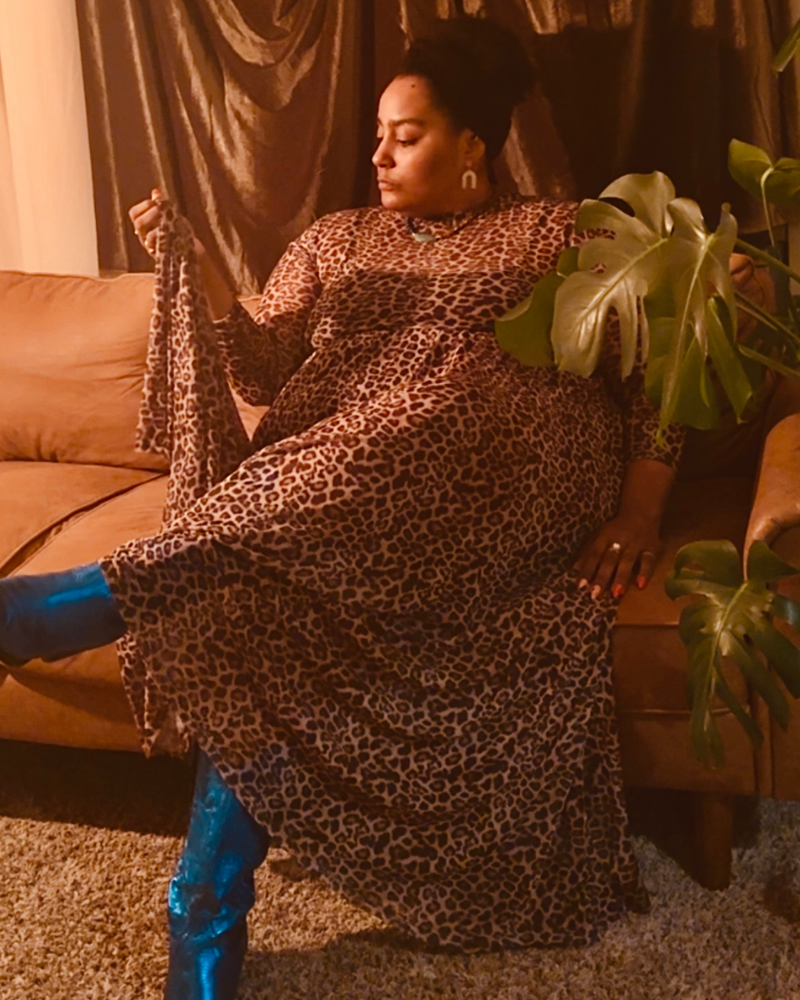 A woman wearing a leopard-print maxi dress and shiny blue boots sits on a brown leather couch in a warm-lit living room with a plant and curtains in the background.