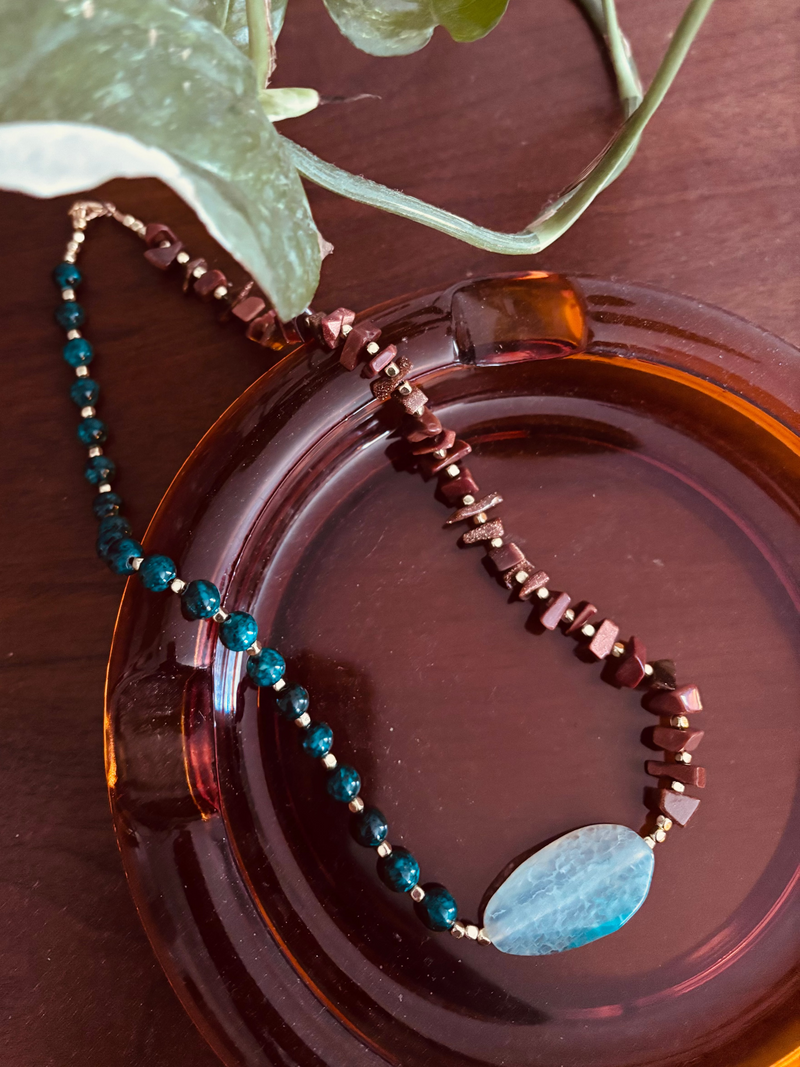 A beaded necklace with dark teal beads, gold-toned spacer beads, and a light blue oval stone pendant, displayed on an amber glass dish with green plant leaves in the background.
