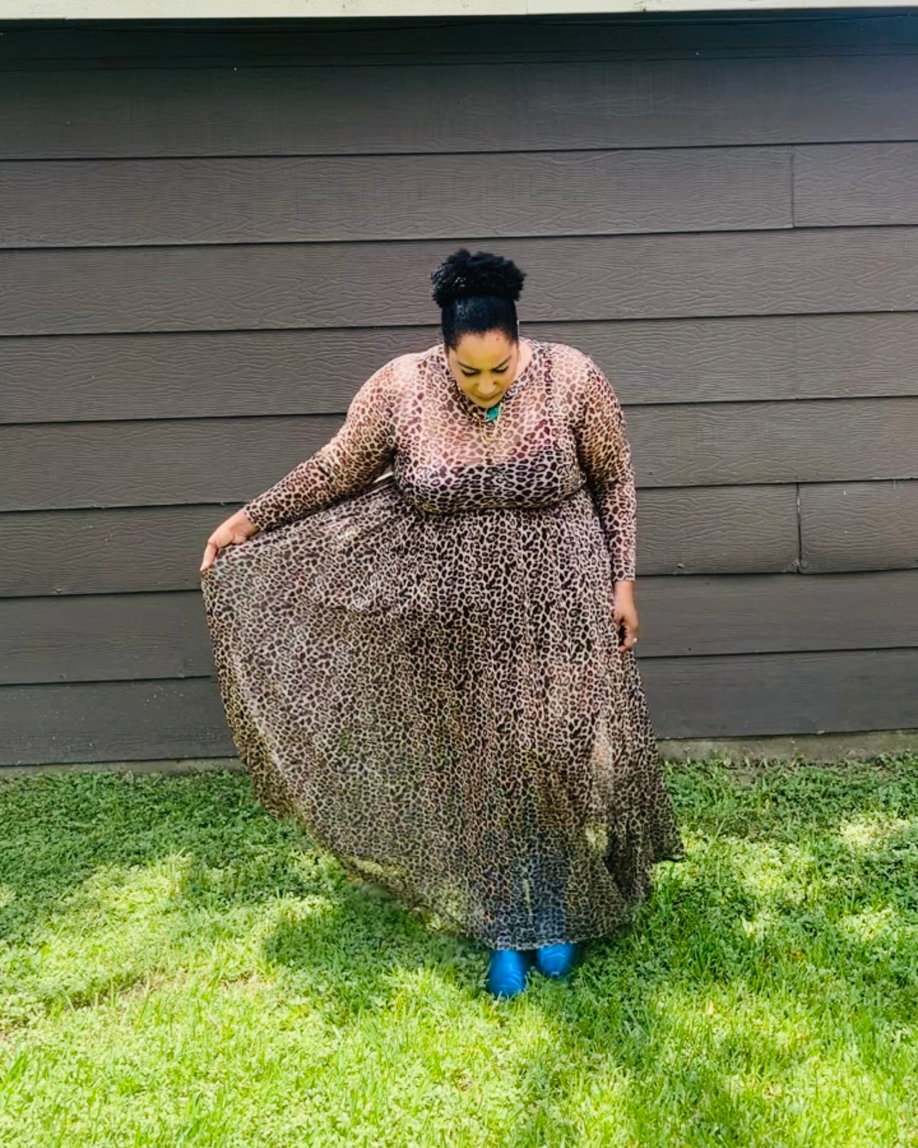 A woman wearing a long-sleeve leopard-print maxi dress and bright blue boots stands on a grassy lawn in front of a dark brown wooden house, holding out the side of the dress to show its full flowy silhouette.