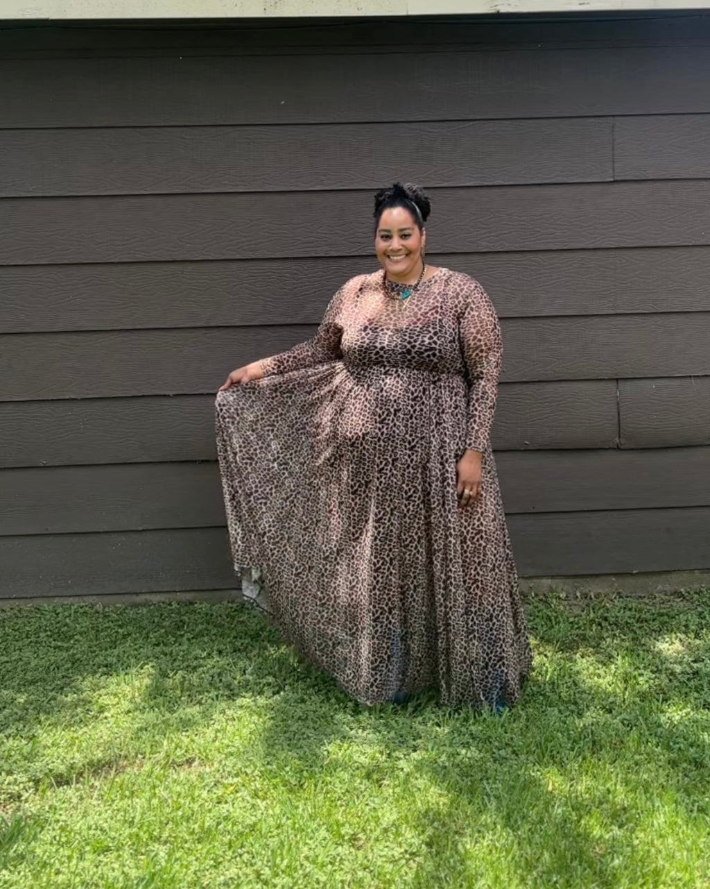 A woman wearing a long-sleeve maxi dress with a brown, black, and beige leopard print stands outdoors in front of a dark gray house wall on a green grassy lawn.