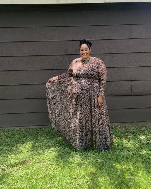 A woman wearing a long-sleeve maxi dress with a brown, black, and beige leopard print stands outdoors in front of a dark gray house wall on a green grassy lawn.