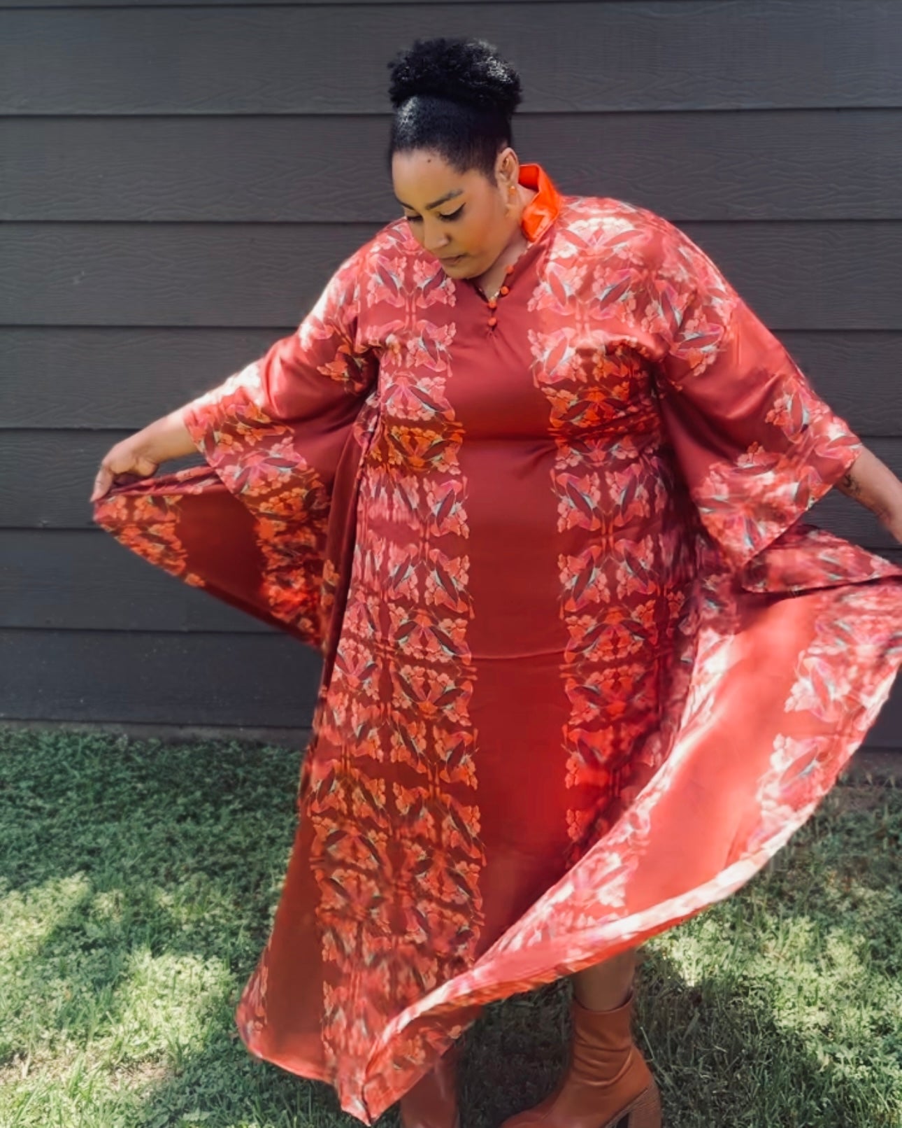 A woman outdoors wearing a flowing wine color kaftan with wide sleeves and a bold geometric pattern, holding the fabric out to show its drape. She is standing in front of a dark gray house siding and green grass.