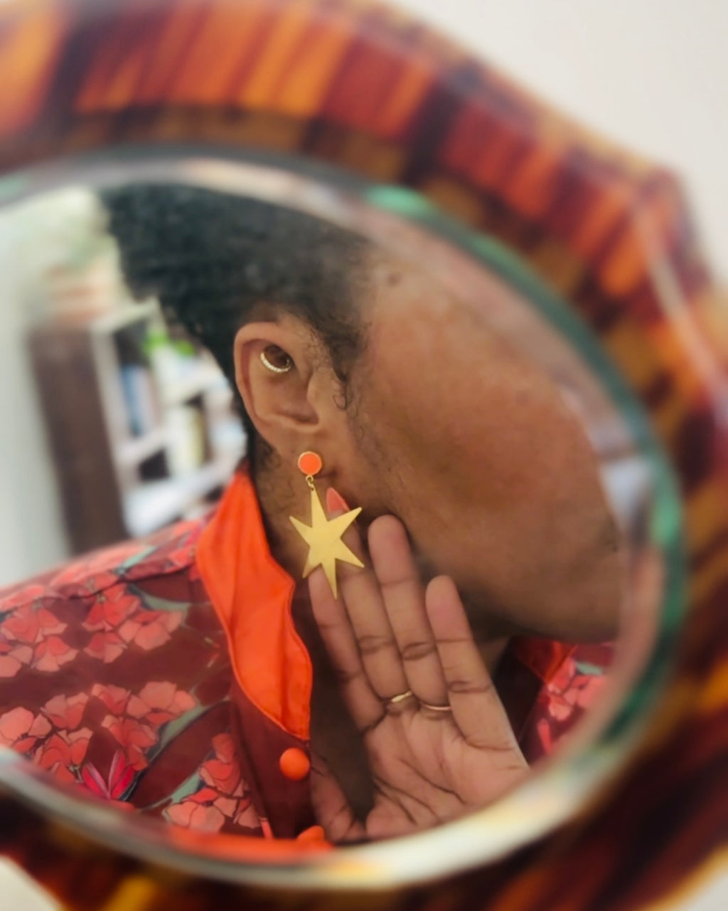 A person wearing a gold star-shaped dangle earring with a small orange bead above it, shown in a close-up mirror reflection. The person is also wearing a red and orange patterned top.