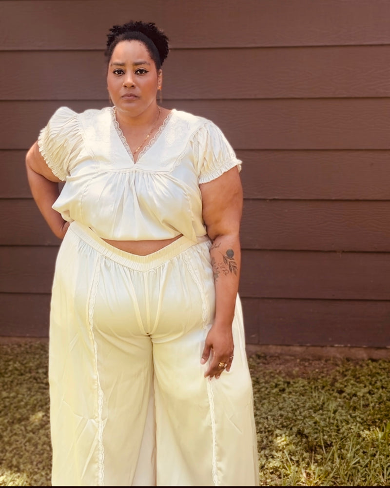 A woman wearing a cream-colored two-piece outfit consisting of a short-sleeve crop top and matching wide-leg pants, standing in front of a brown wooden house exterior.